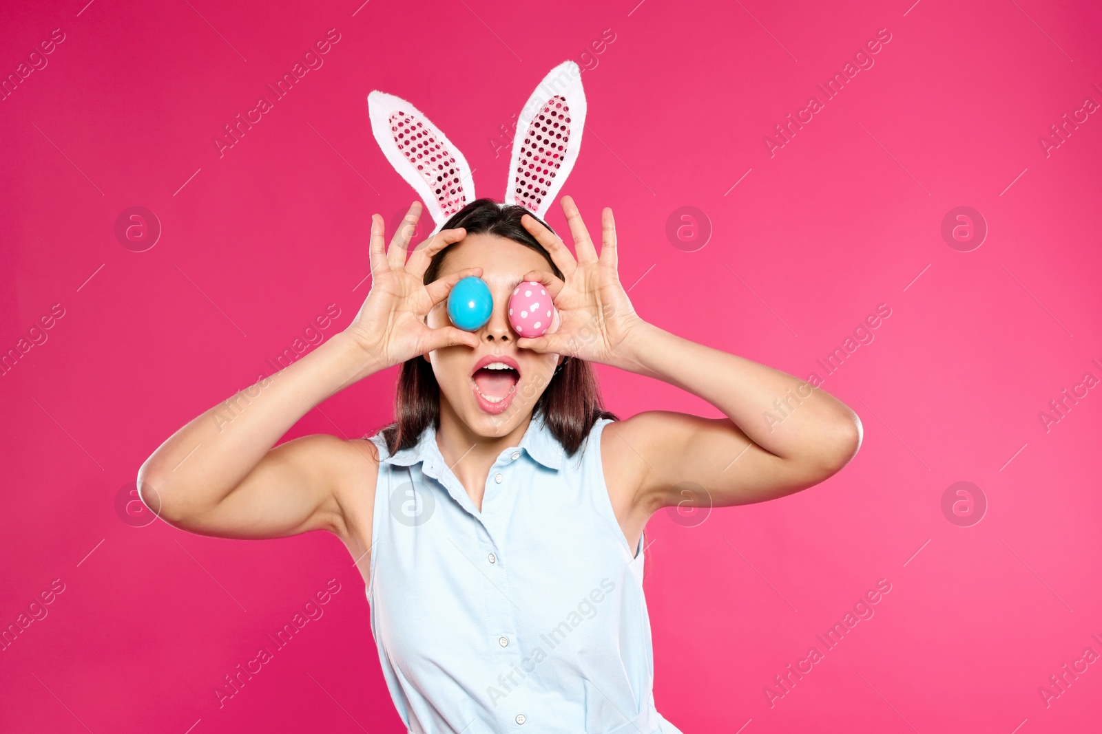 Photo of Beautiful woman in bunny ears headband holding Easter eggs near eyes on color background