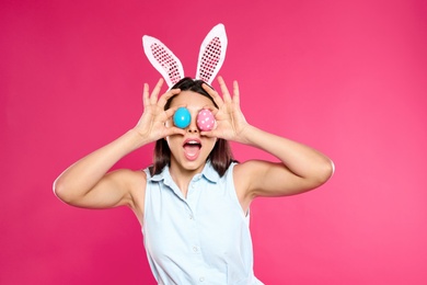 Beautiful woman in bunny ears headband holding Easter eggs near eyes on color background