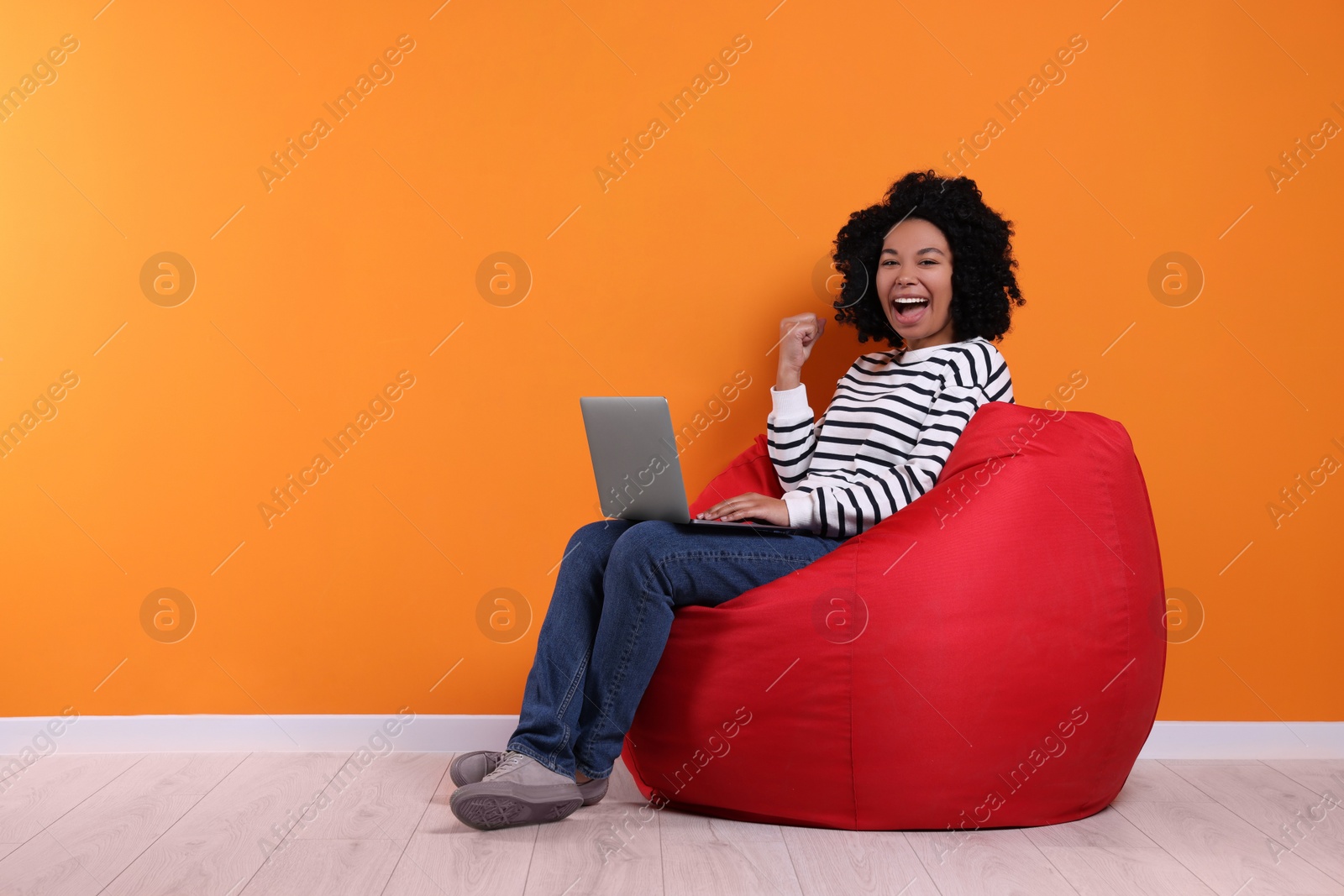 Photo of Emotional young woman with laptop sitting on beanbag chair near orange wall. Space for text