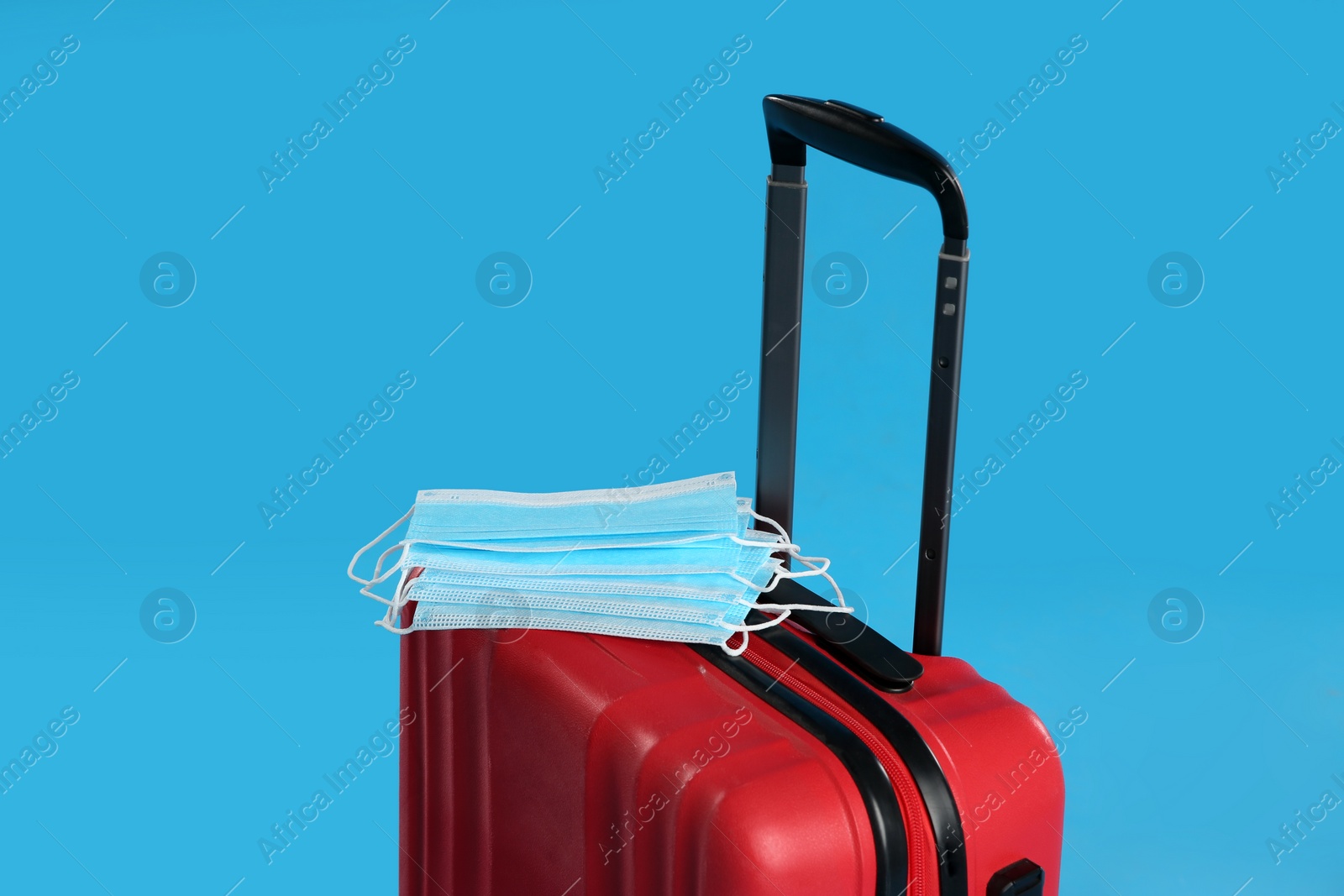 Photo of Stylish red suitcase and protective masks on light blue background. Travelling during coronavirus pandemic