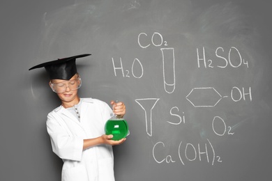 Little school child in laboratory uniform with flask of liquid and chemical formulas on grey background