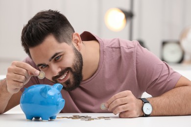 Photo of Happy young man putting money into piggy bank at white table indoors