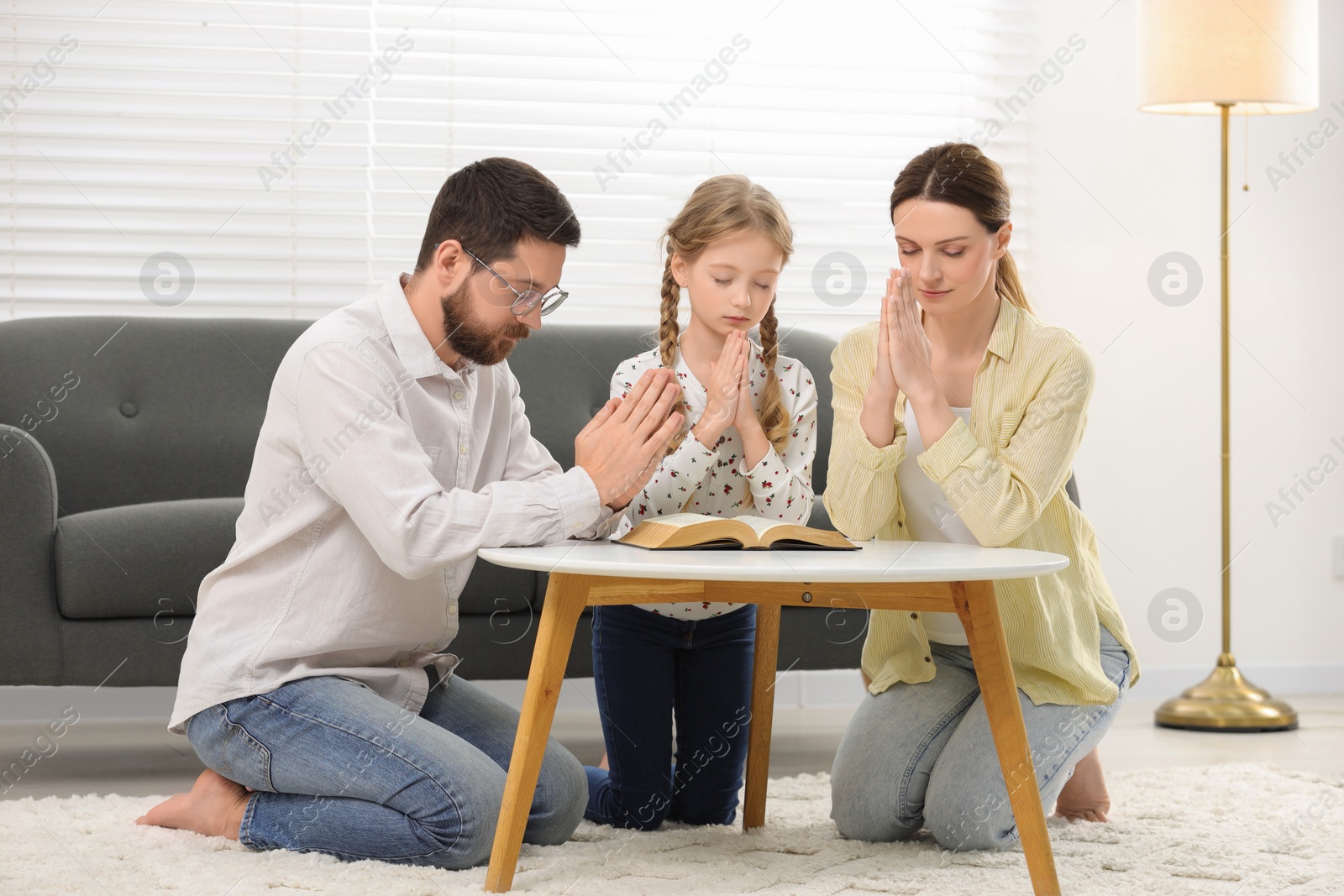Photo of Girl and her godparents praying over Bible together at table indoors