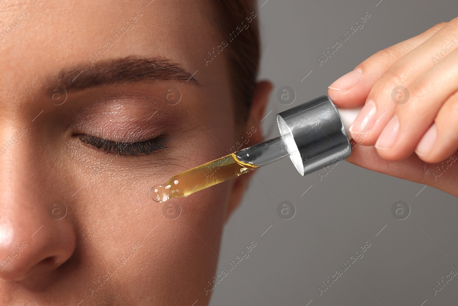 Photo of Beautiful young woman applying cosmetic serum onto her face on grey background, closeup