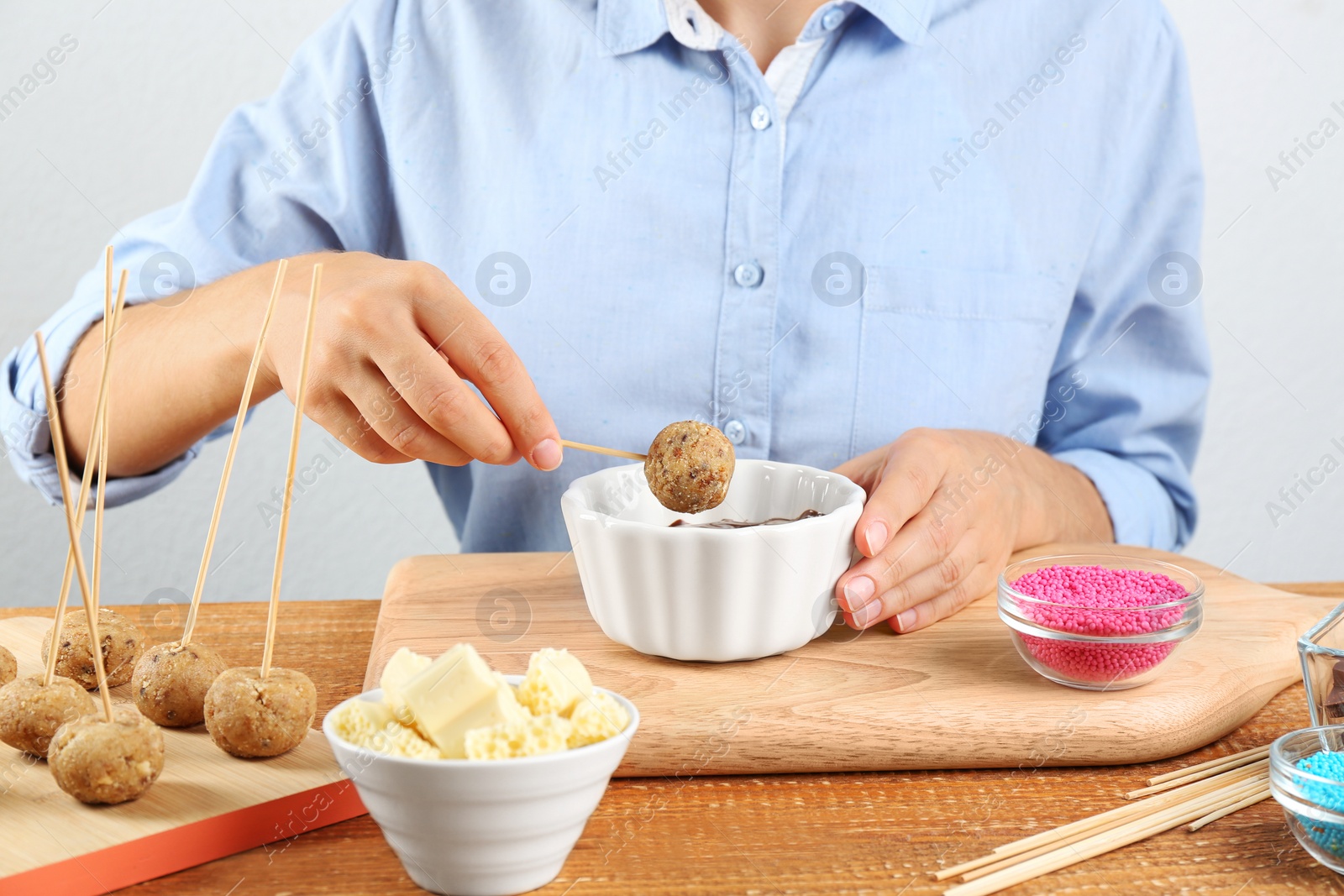 Photo of Young woman putting cake pop into chocolate frosting at wooden table, closeup