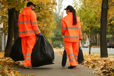 Photo of Street cleaners with brooms and garbage bags outdoors on autumn day, back view