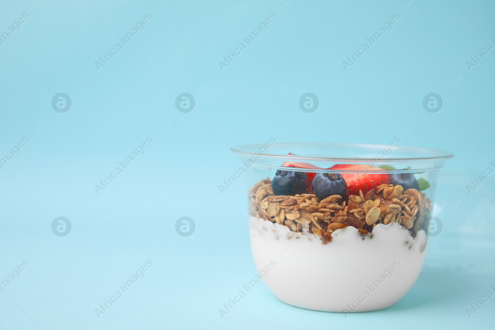 Photo of Tasty granola with berries and yogurt in plastic cup on light blue background, space for text