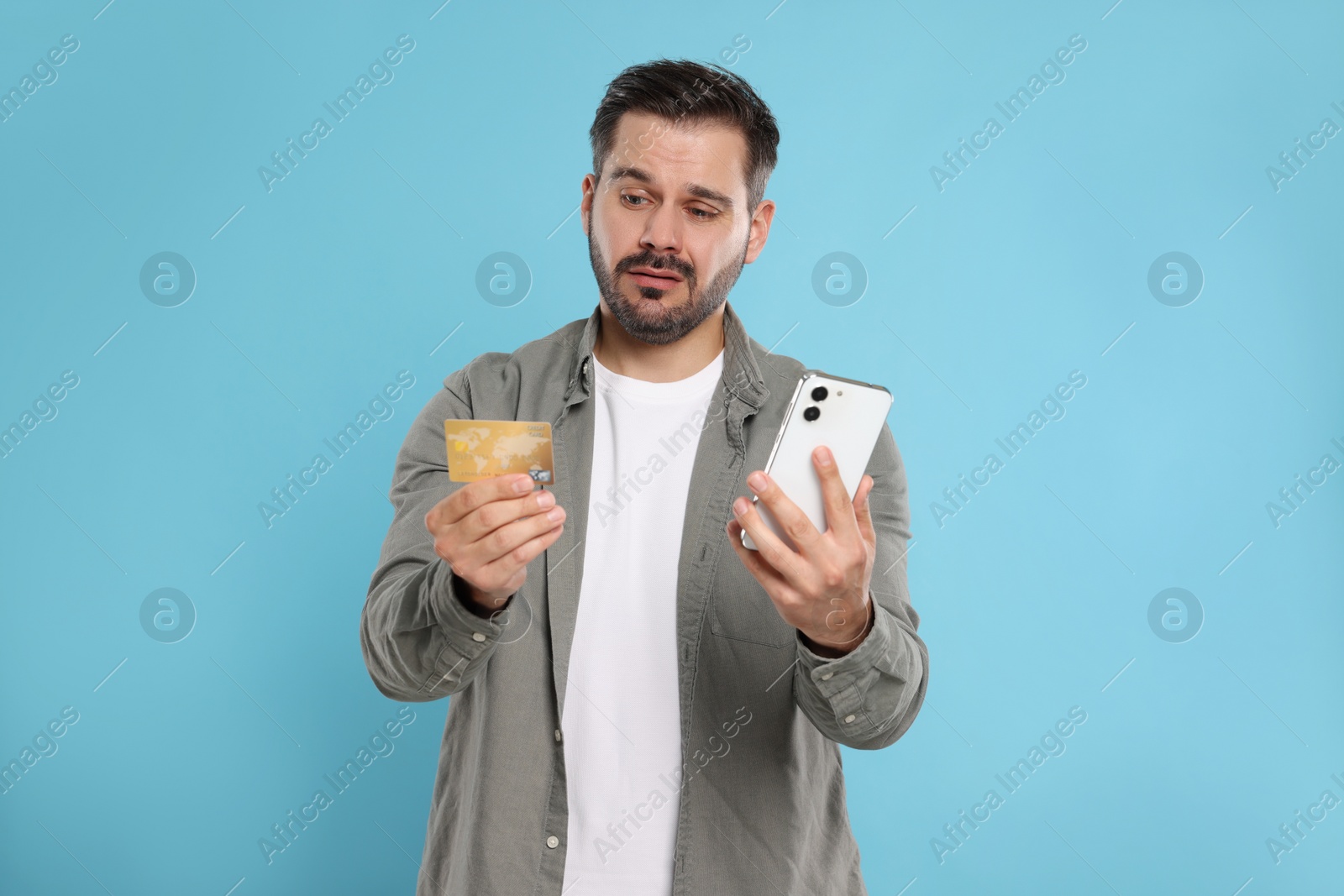 Photo of Confused man with credit card and smartphone on light blue background. Debt problem