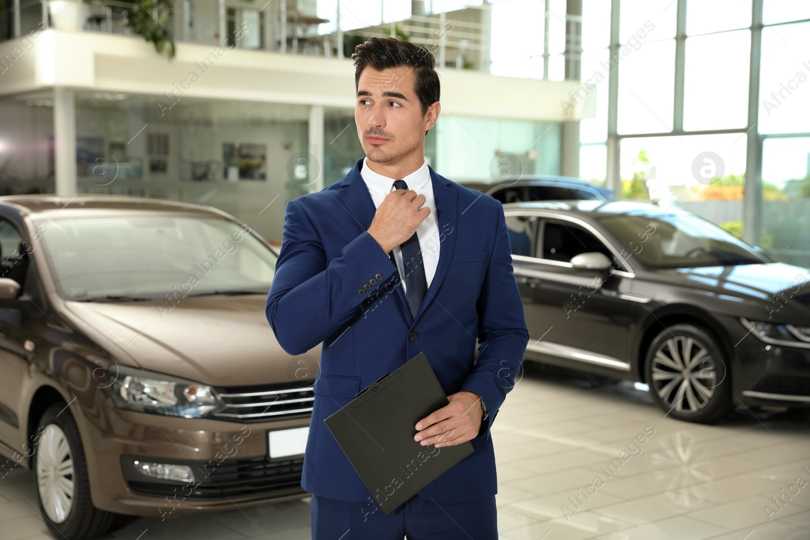 Photo of Young salesman with clipboard near car in modern dealership