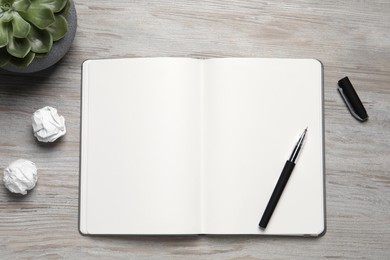 Open blank notebook, plant, stationery and crumpled paper balls on light wooden table, flat lay