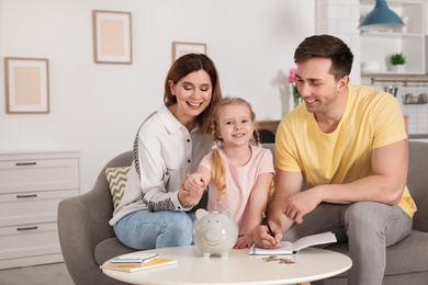 Photo of Happy family putting coin into piggy bank at table indoors. Saving money