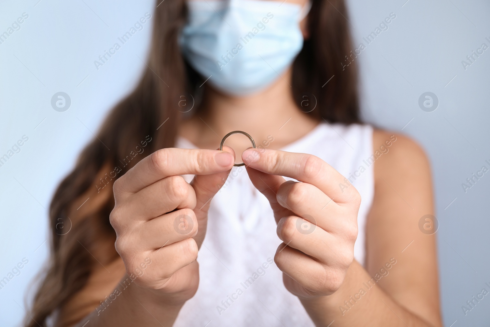 Photo of Woman in protective mask holding wedding ring against light background, focus on hands. Divorce during coronavirus quarantine