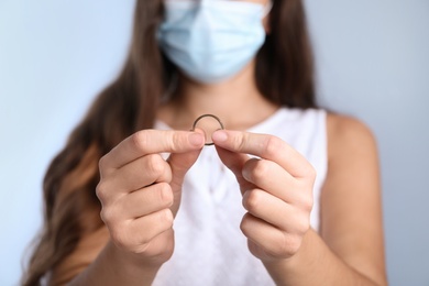 Woman in protective mask holding wedding ring against light background, focus on hands. Divorce during coronavirus quarantine