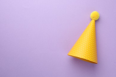 One yellow party hat with pompom on purple background, top view. Space for text