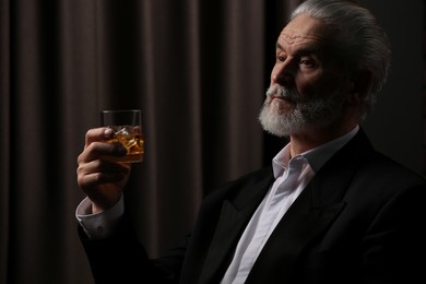 Senior man in formal suit holding glass of whiskey with ice cubes on brown background