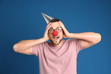 Photo of Emotional man with glasses, party hat and clown nose on blue background. April fool's day