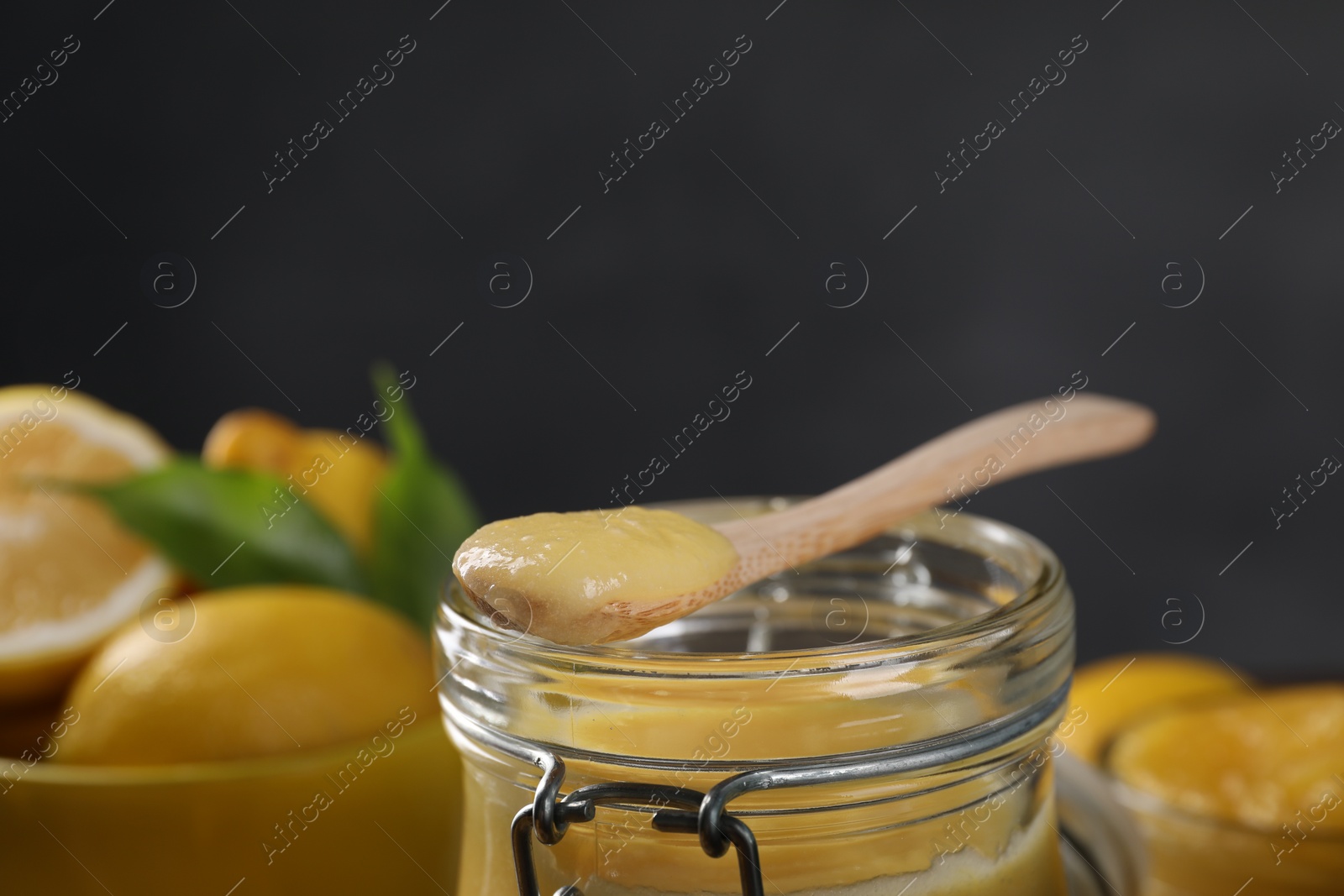 Photo of Delicious lemon curd in glass jar, spoon and fresh citrus fruits against blurred background, closeup