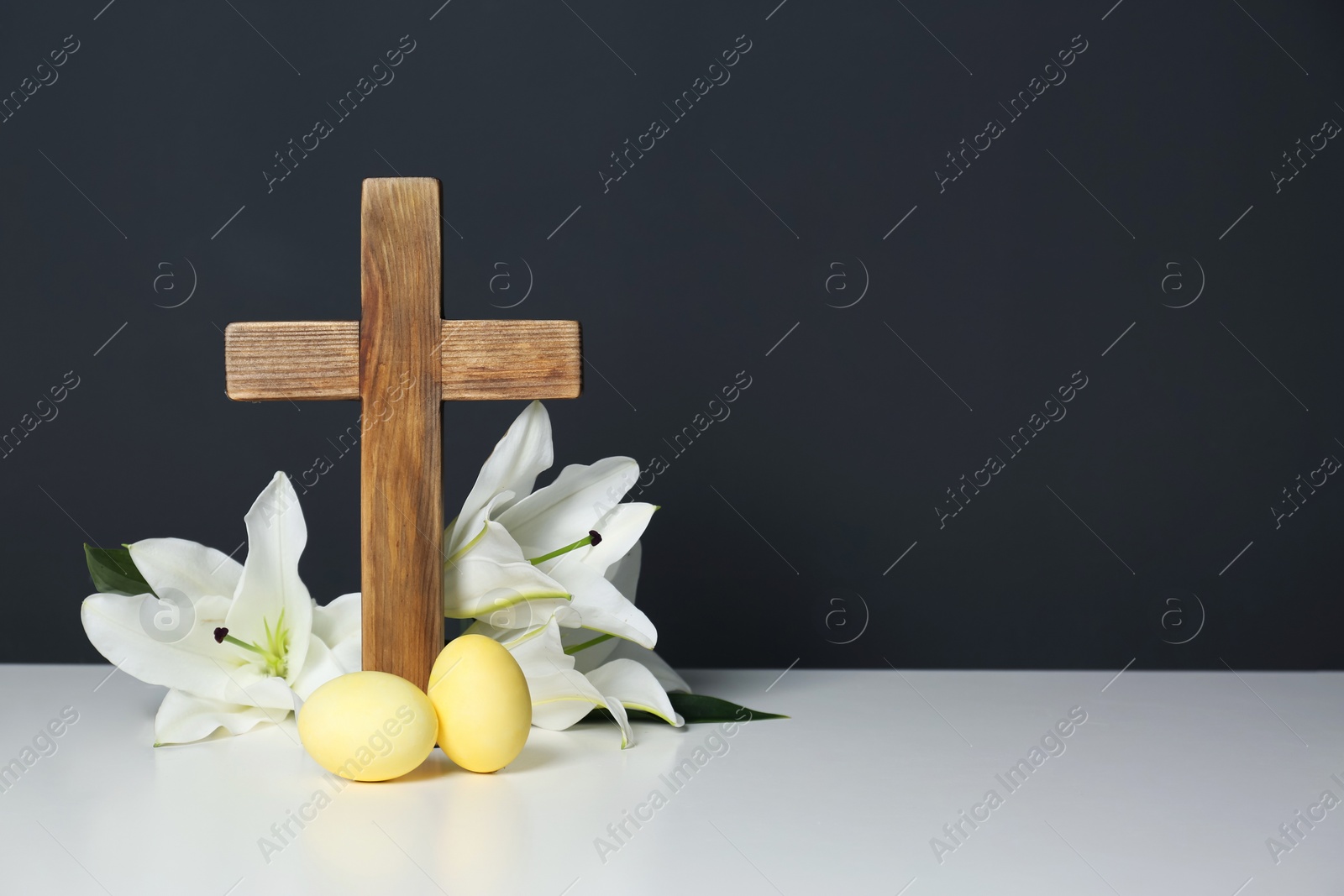 Photo of Wooden cross, Easter eggs and blossom lilies on table against color background, space for text