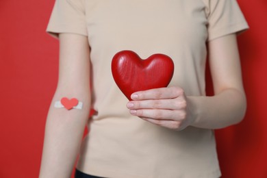 Photo of Blood donation concept. Woman with adhesive plaster on arm holding red heart against color background, closeup