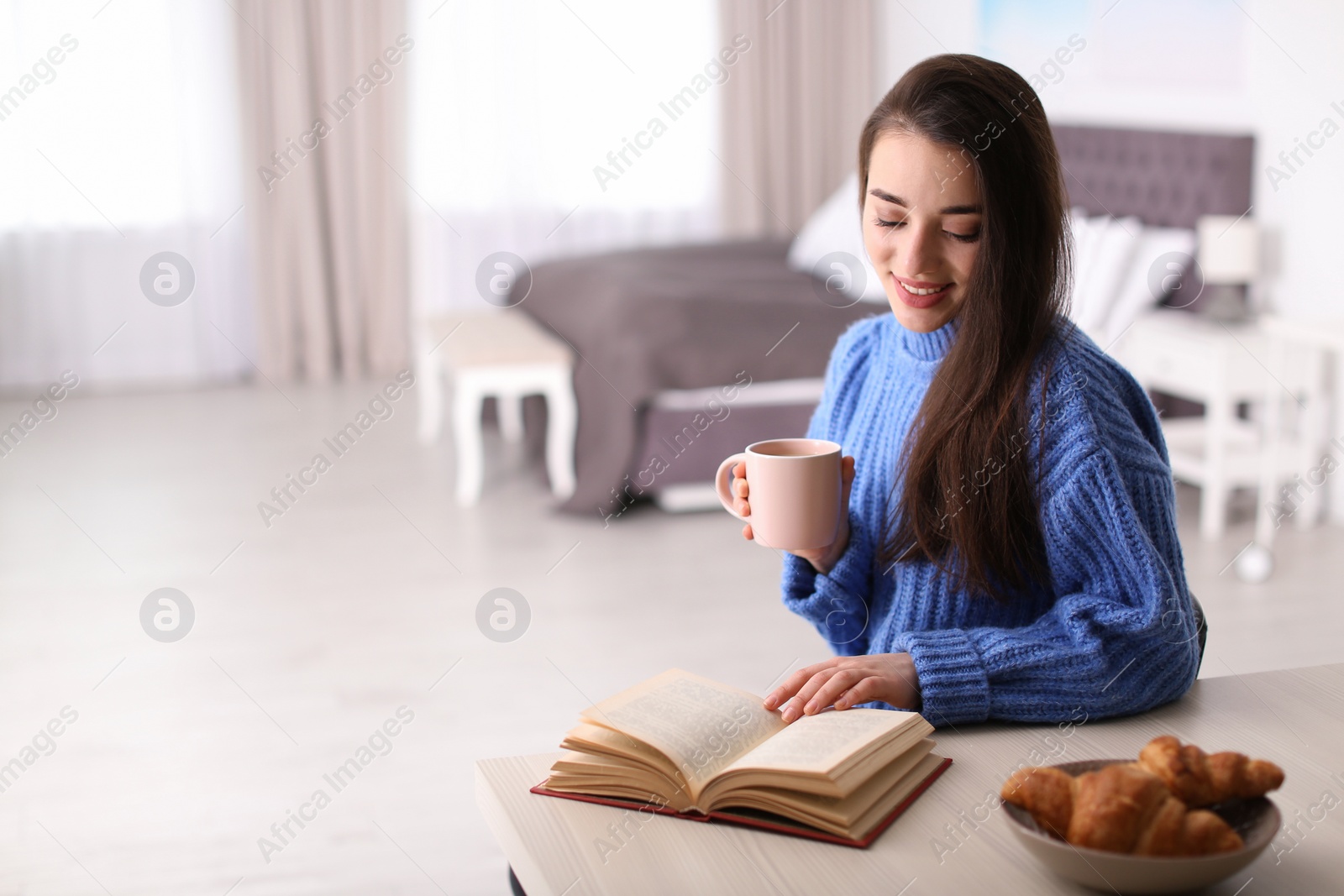 Photo of Young woman drinking coffee and reading book at table indoors, space for text. Winter season