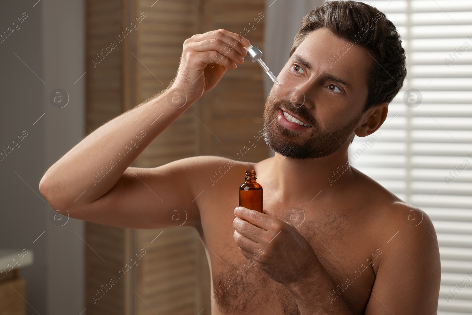 Photo of Smiling man applying cosmetic serum onto his face indoors