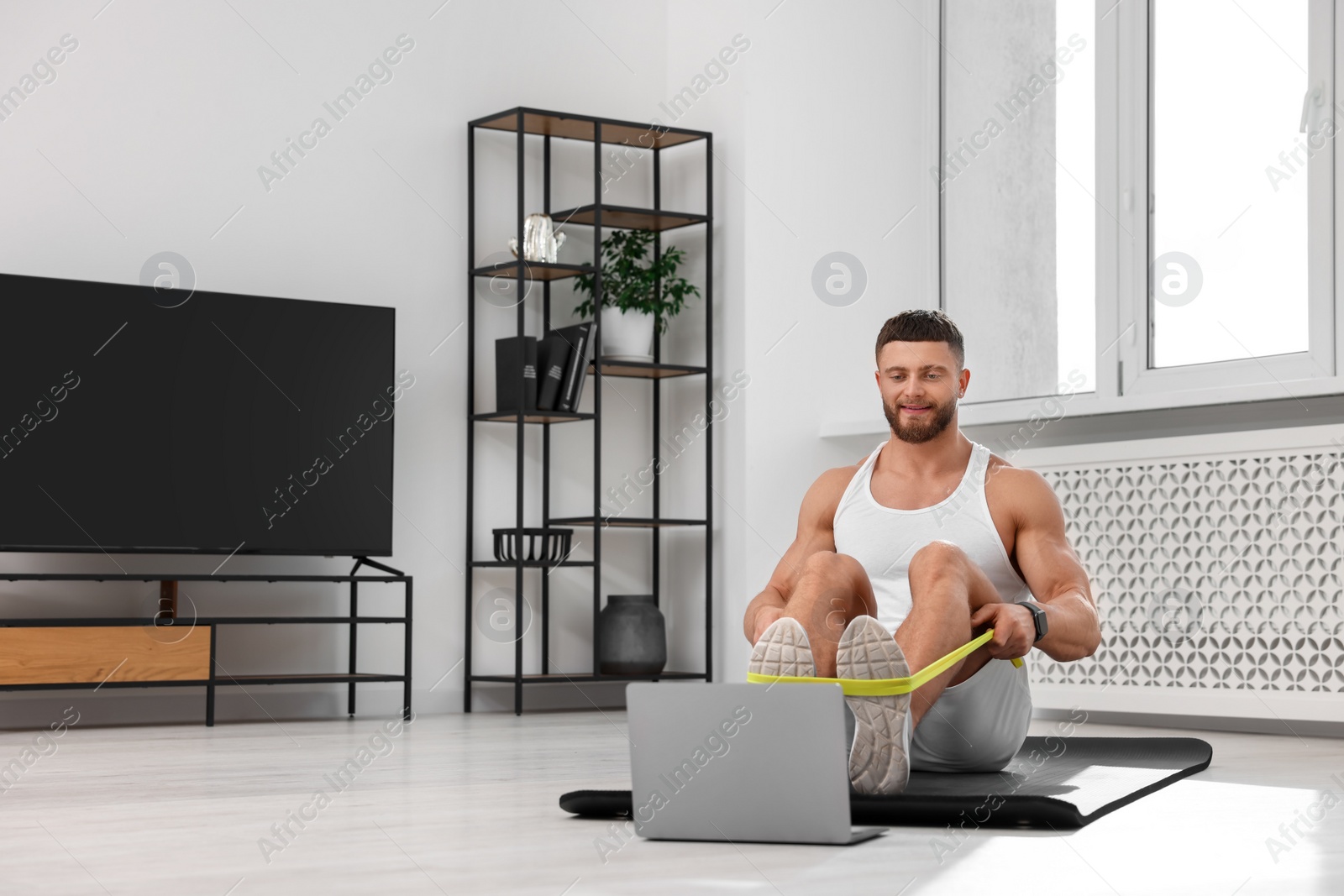 Photo of Muscular man doing exercise with elastic resistance band near laptop on mat at home