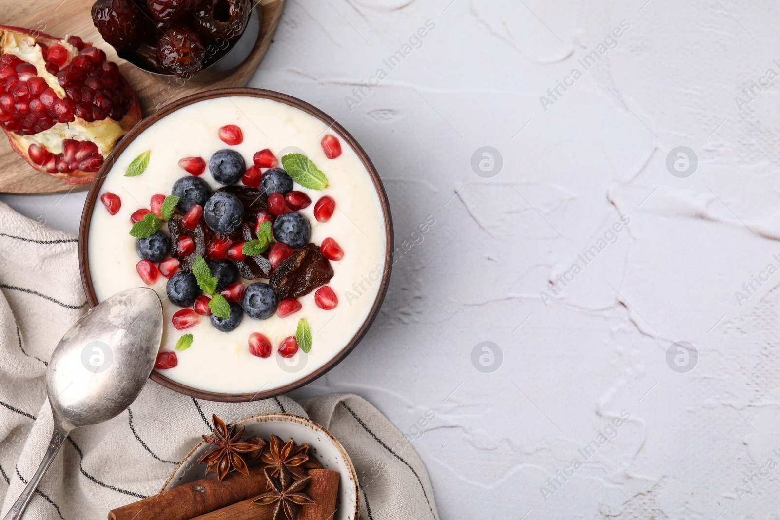Photo of Delicious semolina pudding with blueberries, pomegranate, dates and mint in bowl served on white textured table, top view. Space for text