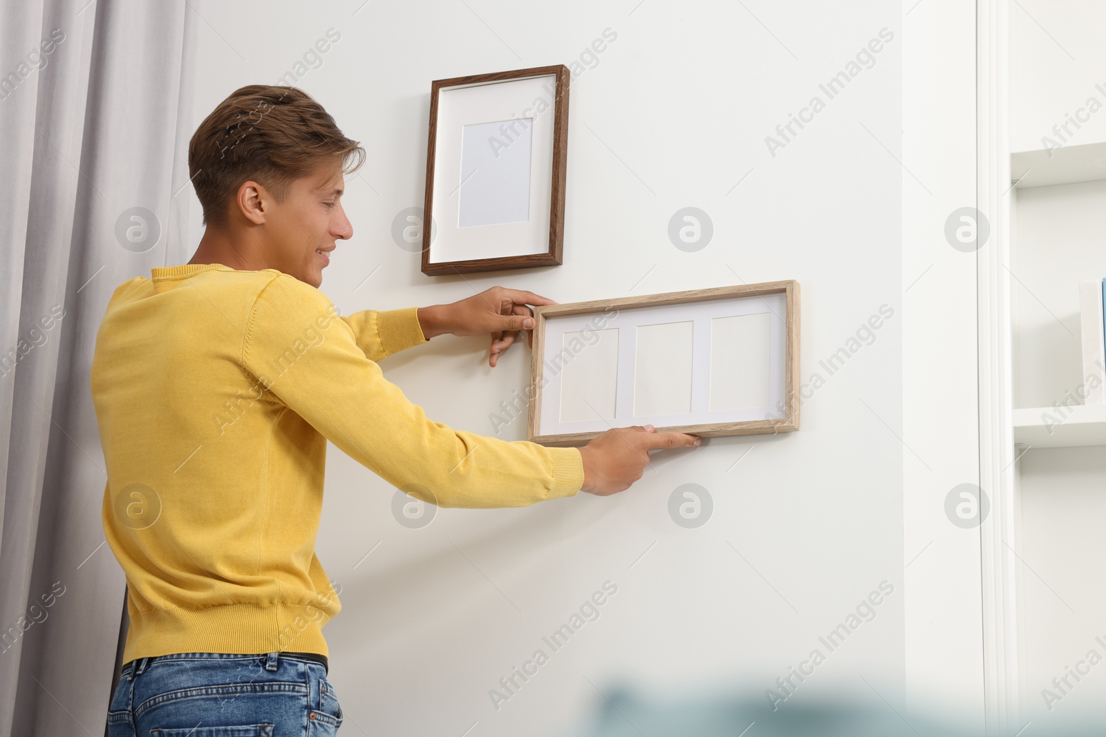 Photo of Young man hanging picture frames on white wall indoors