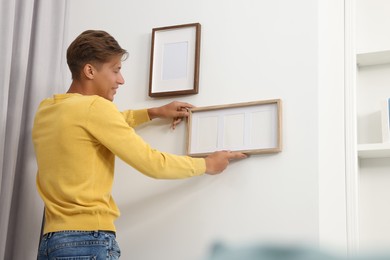 Young man hanging picture frames on white wall indoors