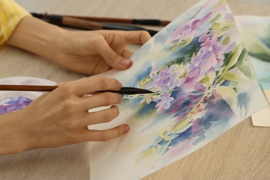 Photo of Woman painting flowers with watercolor at white wooden table, closeup. Creative artwork