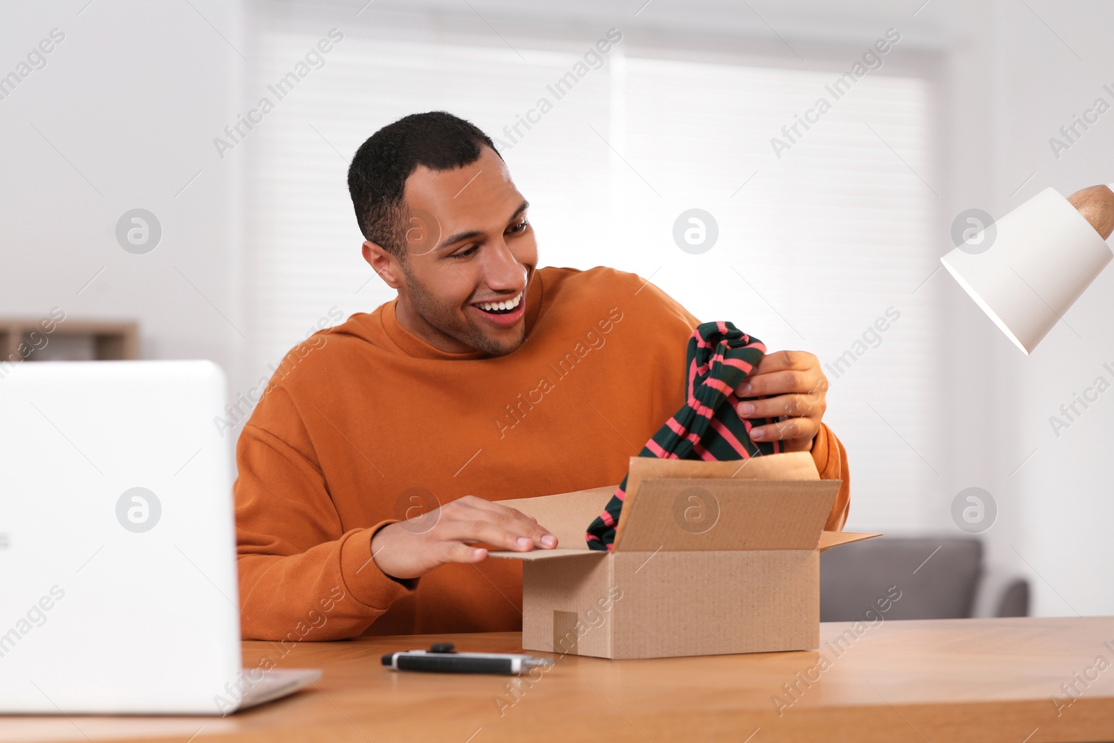 Photo of Happy young man opening parcel at table indoors. Internet shopping