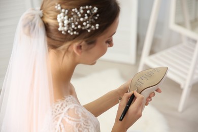 Young bride writing her single friends names on shoe indoors, closeup. Wedding superstition