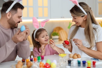 Photo of Easter celebration. Happy family with bunny ears painting eggs at white marble table in kitchen