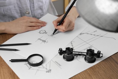 Jeweler drawing sketch of elegant ring on paper at wooden table, closeup