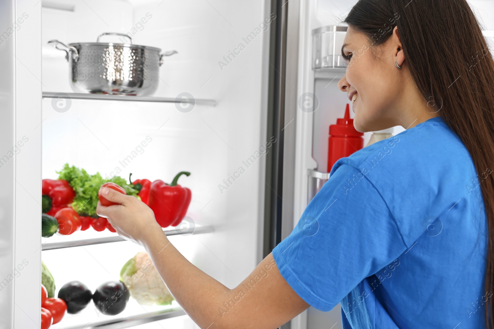 Photo of Young woman taking tomato out of refrigerator
