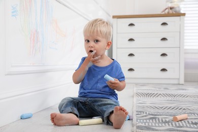 Mischievous little boy near white wall with colorful chalk scribbles at home