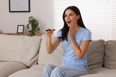 Emotional woman with remote controller watching TV on sofa at home