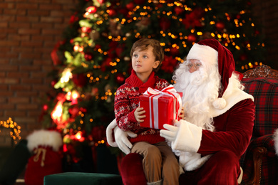 Photo of Santa Claus and little boy with gift near Christmas tree indoors