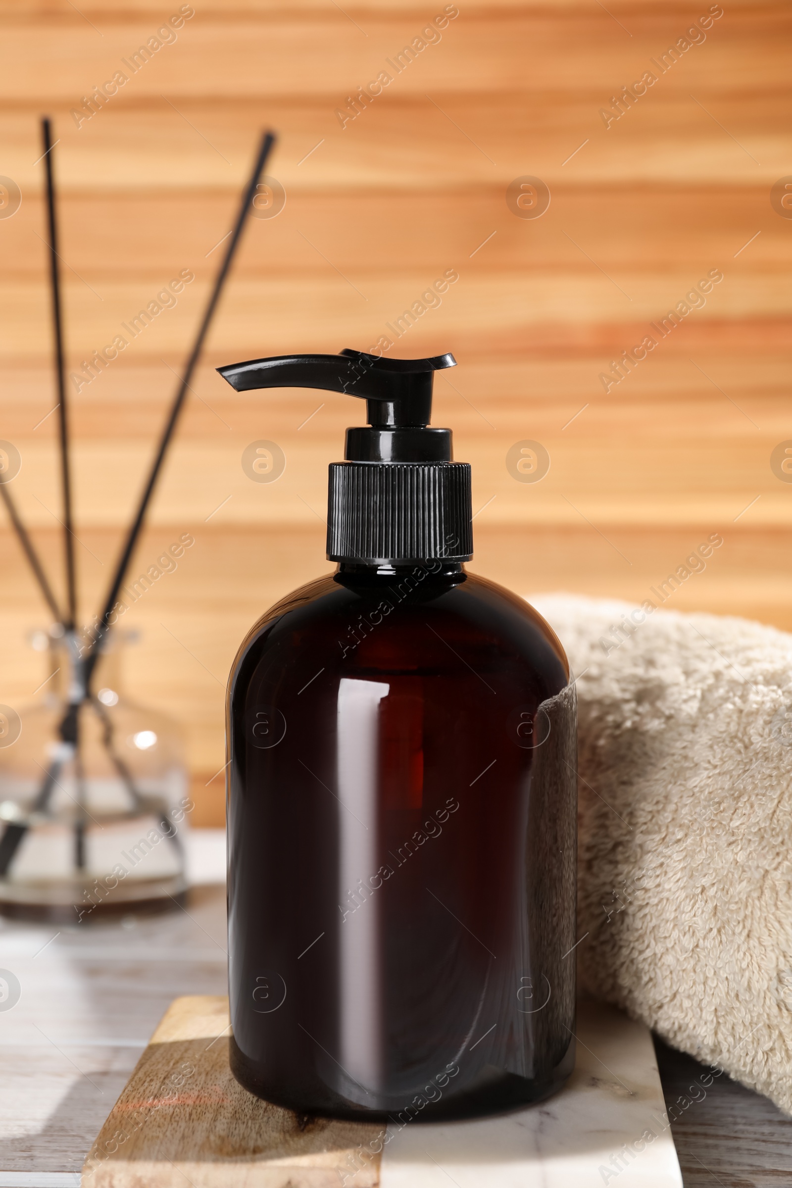 Photo of Bottle of shampoo and rolled towel on white table near wooden wall, closeup