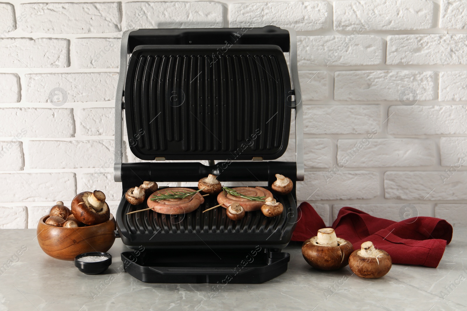 Photo of Electric grill with homemade sausages, rosemary and mushrooms on marble table