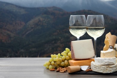 Different types of delicious cheeses, snacks and wine on wooden table against mountain landscape. Space for text