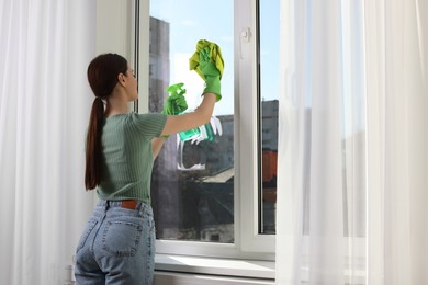Photo of Young woman cleaning window glass with rag and detergent at home, space for text
