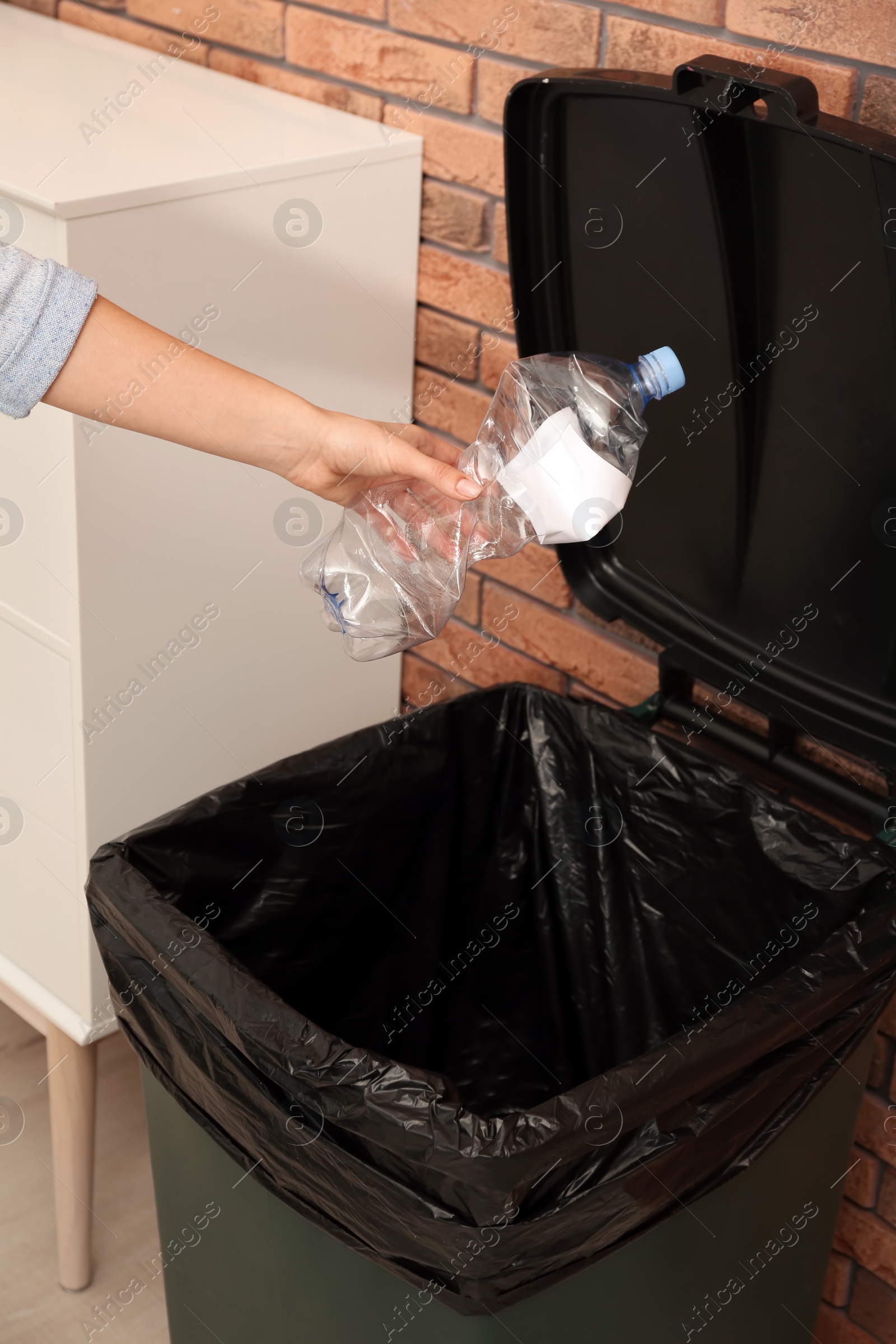 Photo of Young woman throwing empty plastic bottle in trash bin indoors, closeup. Waste recycling