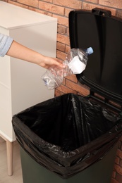 Young woman throwing empty plastic bottle in trash bin indoors, closeup. Waste recycling