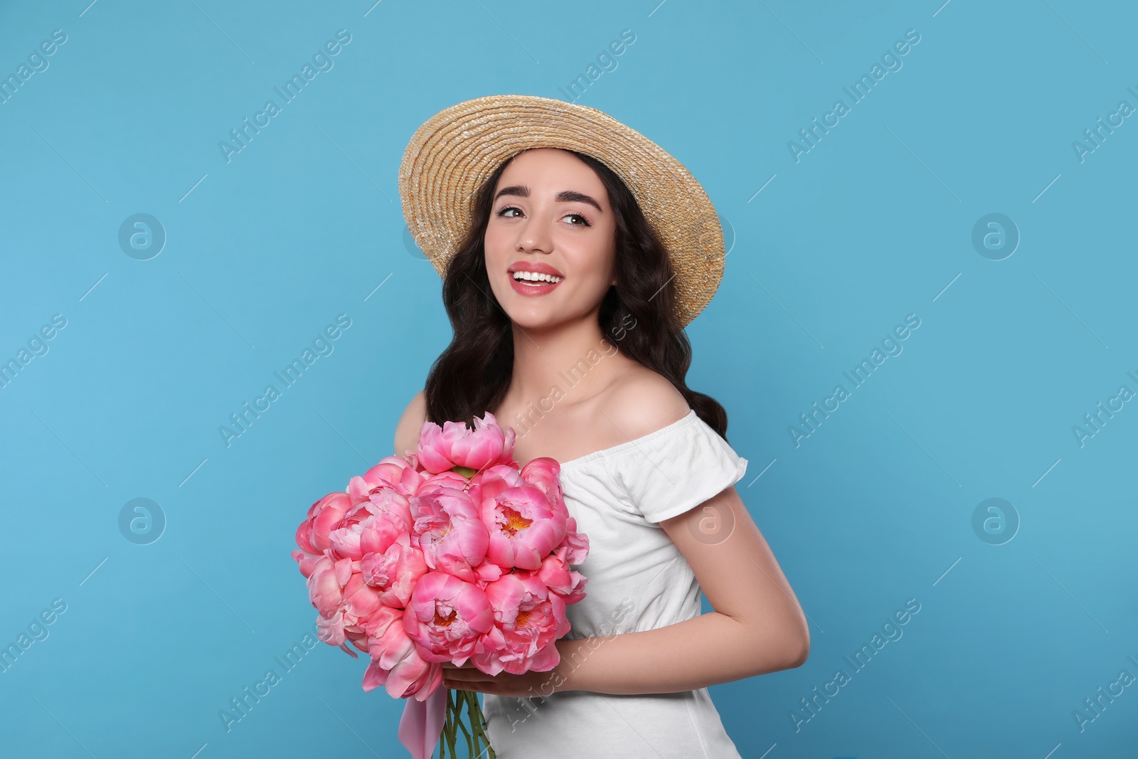 Photo of Beautiful young woman in straw hat with bouquet of pink peonies against light blue background