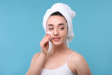 Beautiful woman in terry towel removing makeup with cotton pad on light blue background