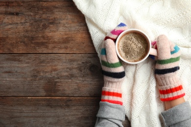 Woman with knitted mittens holding hot winter drink on wooden background, top view. Space for text