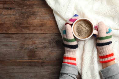 Photo of Woman with knitted mittens holding hot winter drink on wooden background, top view. Space for text