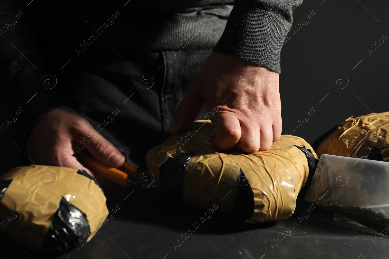 Photo of Smuggling and drug trafficking. Man opening package of narcotics with box cutter at dark table, closeup