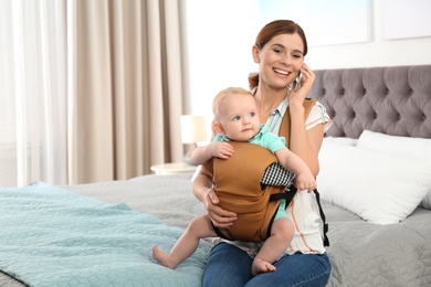 Photo of Woman with her son in baby carrier talking on phone at home. Space for text